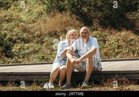 Porträt eines glücklichen älteren Ehepaares, das auf einem Holzweg in einem Kiefernwald in den Dünen sitzt und spricht. Stockfoto