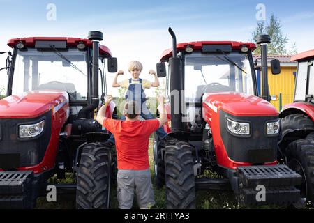 Mann und Junge stehen vor großen roten neuen Traktoren am Verkaufsstandort. Ein Erwachsener und ein Kind sehen sich landwirtschaftliche Maschinen mit Interesse an und zeigen ihre Kraft Stockfoto