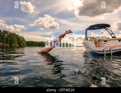 Mann, der am Sommertag vom Boot in einen See taucht, während der Hund beobachtet. Stockfoto