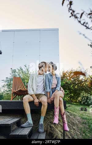 Multiethnisches Paar in stilvoller Freizeitkleidung, das sich auf der Veranda eines Glashauses auf dem Land küsst Stockfoto