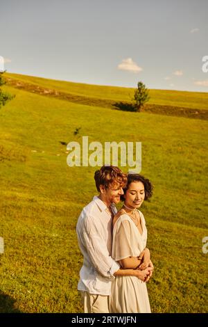 Gerade verheiratetes Paar, glücklicher Mann, der asiatische Braut umarmt, im grünen Feld, landschaftlich reizvolle und ruhige Landschaft Stockfoto