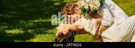 Junger rothaariger Mann umarmt und küsst asiatische Frau mit Hochzeitsstrauß im Freien, Banner Stockfoto
