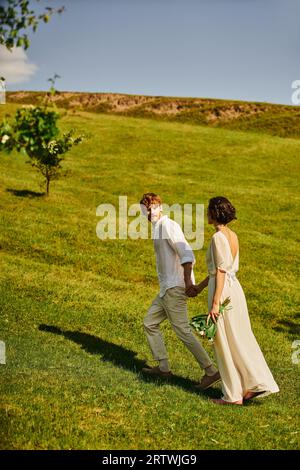 Ich habe gerade ein interrassisches Paar in weißer Hochzeitskleidung geheiratet, das die Hände hält und auf einer grünen Wiese läuft Stockfoto