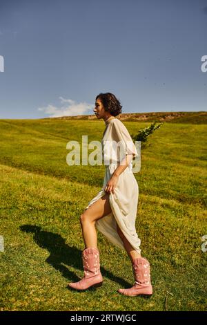 Seitenansicht einer jungen asiatischen Frau in Brautkleidern und Cowboystiefeln, die auf einer Wiese auf dem Land spazieren Stockfoto