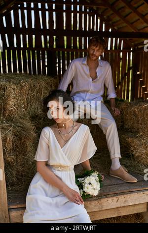 asiatische Braut in weißem Kleid, die Blumenstrauß hält und auf Heu neben rotem Bräutigam in Scheune sitzt, ländlich Stockfoto