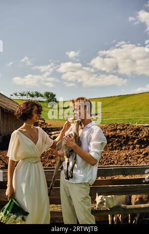 Freudvolles interrassisches Brautpaar, das süße Ziegenbabys kuschelt, asiatische Braut in Hochzeitskleid und Sonnenbrille Stockfoto