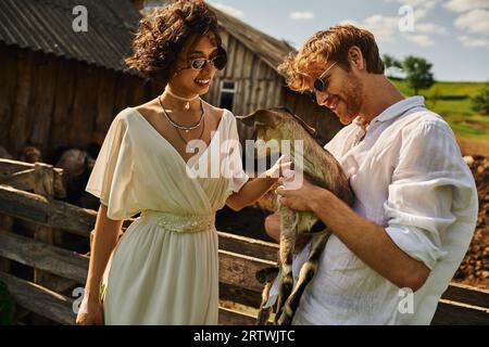 Fröhliche, multiethnische Brautpaare, die süße Ziegenbabys, eine asiatische Braut im Hochzeitskleid und in der Sonnenbrille kuscheln Stockfoto