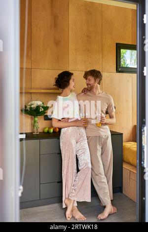 Fröhliches Paar, das barfuß im Pyjama steht und morgens Gläser mit Orangensaft hält Stockfoto