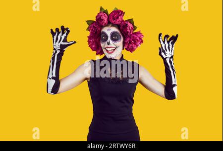 Mädchen mit Zuckerschädel-Make-up, das mit den Händen Angst macht und auf gelbem Hintergrund lächelt. Stockfoto