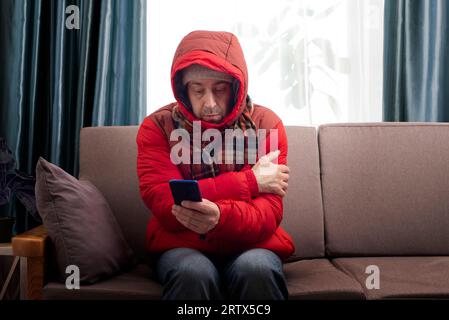 Männliches Modell mittleren Alters in rotem Parka und Schal, das auf dem Sofa sitzt, während es sein Smartphone in seinem kalten Wohnzimmer benutzt Stockfoto