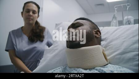 Junge Krankenschwester bringt verletzten Mann mittleren Alters auf Krankenhausstation. Der afroamerikanische Patient liegt auf einer trage in der Nackenstütze und spricht mit dem Arzt über die Gesundheit. Medizinisches Personal am Arbeitsplatz im medizinischen Zentrum. Stockfoto