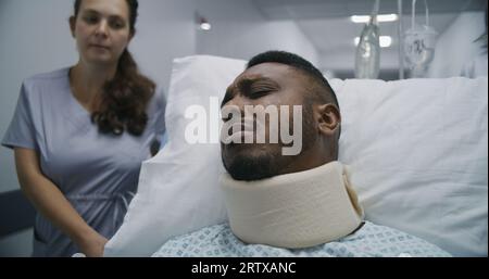 Junge Krankenschwester bringt verletzten Mann mittleren Alters auf Krankenhausstation. Der afroamerikanische Patient liegt auf einer trage in der Nackenstütze und spricht mit dem Arzt über die Gesundheit. Medizinisches Personal am Arbeitsplatz im medizinischen Zentrum. Stockfoto