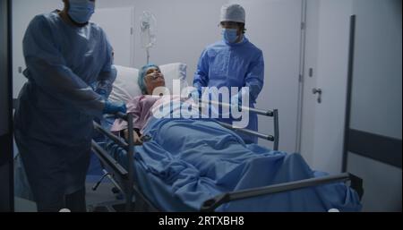 Ärzte, Pflegekräfte und Sanitäter schieben die Krankenstation mit dem kranken Patienten in den Operationssaal. Ältere Frau liegt auf einer trage mit medizinischem Tropf. Personalarbeit in der Notaufnahme. Rückansicht. Die Aufnahme wird verfolgt. Stockfoto