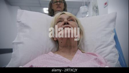 Ältere Frau liegt auf der Bahre, spricht mit reifer Ärztin. Der Arzt bringt den Patienten nach der Operation auf die Krankenhausstation in den modernen Flur des medizinischen Zentrums. Medizinisches Personal am Arbeitsplatz im Krankenhaus. Stockfoto