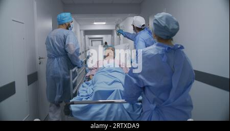 Ärzte, Pflegekräfte und Sanitäter schieben die Krankenstation mit dem kranken Patienten in den Operationssaal. Ältere Frau liegt auf einer trage mit medizinischem Tropf. Personalarbeit in der Notaufnahme. Rückansicht. Die Aufnahme wird verfolgt. Stockfoto