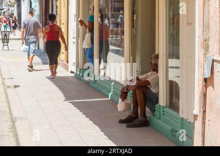 Havanna, Kuba, 2023, Ein kubanischer Mann sitzt vor der Tür eines Gebäudes. Andere Leute sind auf dem Bürgersteig unterwegs. Stockfoto