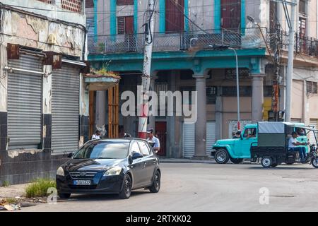 Havanna, Kuba, 2023: Ein modernes Auto steht im Kontrast zu alten Fahrzeugen in einer Stadtstraße – die traditionellen heruntergekommenen und verwitterten Gebäude stehen im Hintergrund. Stockfoto