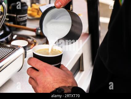 Barista bereitet Kaffee zu. Gießen Sie Milch aus dem Becher in eine Tasse Kaffee, der von einem Barista zubereitet wird. Konzentriere dich auf die Hände, die eine Tasse Kaffee halten Stockfoto