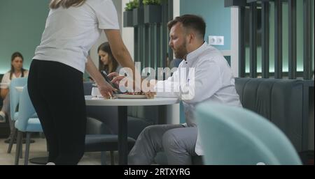 Erwachsener Arzt arbeitet auf digitalem Tablet und isst sein Abendessen im Krankenhaus Café. Cafeteria Arbeiter bringt Essen für Sanitäter. Die Patientin verwendet ihr Telefon im Hintergrund. Medizinisches Personal hat Pause in der Klinikkantine. Stockfoto