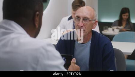 Ein älterer Patient sitzt mit einem afroamerikanischen Arzt im Klinikcafé. Professioneller Arzt diskutiert medizinische Testergebnisse mit dem Menschen. Digitales Tablet mit MRT-Scan-Bild. Krankenhaus oder medizinisches Zentrum Cafeteria. Stockfoto