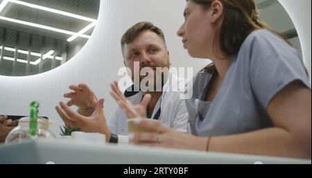 Männlicher Arzt und Krankenschwester sitzen in einem modernen Klinikcafé. Professionelle Sanitäter essen Essen, unterhalten sich beim Abendessen, diskutieren über Arbeit. Medizinisches Personal im Krankenhaus oder medizinische Zentrum Cafeteria. Lifestyle- und Gesundheitskonzept. Stockfoto