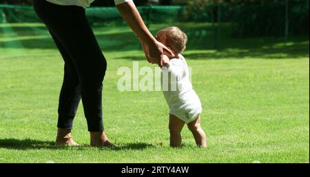 Das süße Baby lehnt sich draußen auf die Beine der Mutter. Entzückendes Kleinkind, das neben der Mutter steht und sich im Freien aufhält Stockfoto