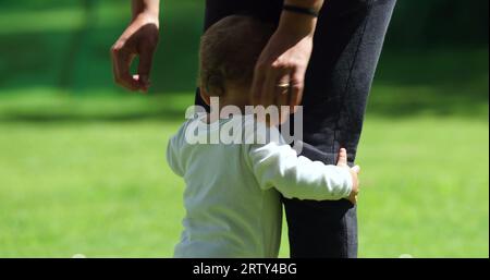 Das süße Baby lehnt sich draußen auf die Beine der Mutter. Entzückendes Kleinkind, das neben der Mutter steht und sich im Freien aufhält Stockfoto