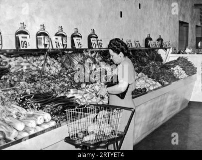 Vereinigte Staaten um 1940. Eine Frau in einem Lebensmittelgeschäft, die eine Auswahl aus der Gemüseabteilung macht. Sie verwendet einen frühen Warenkorb, der den separaten Korb trägt und zusammenklappt, wenn er nicht verwendet wird. Stockfoto