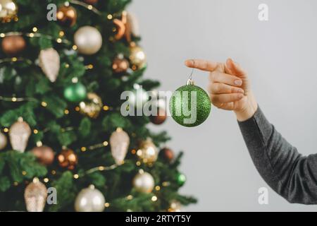 Frau, die den großen Weihnachtsbaum dekoriert. Weibchen mittleren Alters, das eine grüne Kugel in der Hand hält und am Baumzweig hängt. Leerzeichen kopieren. Grußkarte. Nahaufnahme Stockfoto