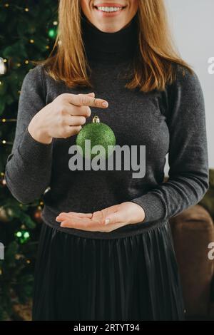Frau, die den großen Weihnachtsbaum dekoriert. Weibchen mittleren Alters, das eine grüne Kugel in der Hand hält und am Baumzweig hängt. Leerzeichen kopieren. Grußkarte. Nahaufnahme Stockfoto