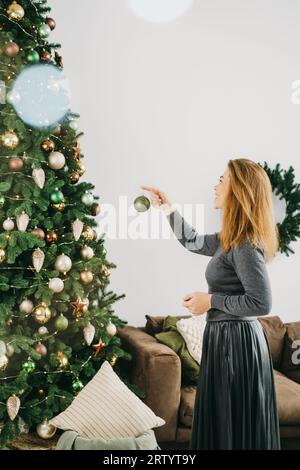 Frau, die den großen Weihnachtsbaum dekoriert. Weibchen mittleren Alters, das eine grüne Kugel in der Hand hält und am Baumzweig hängt. Leerzeichen kopieren. Grußkarte. Stockfoto