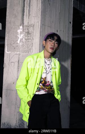 Ein asiatischer Mann in einer kalkfarbenen Jacke, die sich an einer stillgelegten Gebäudesäule lehnt und nachts sehr hübsch aussieht Stockfoto