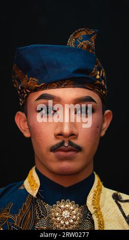 Ein asiatischer Mann, der ein traditionelles indonesisches Tanzkostüm trägt, ist braun und sein Gesicht ist voller Make-up in einem Raum Stockfoto