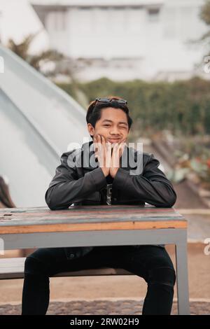 Ein asiatischer Mann mit einem molligen Gesicht, der eine Sonnenbrille und eine schwarze Lederjacke trägt, während er morgens an einem Kaffeetisch sitzt Stockfoto