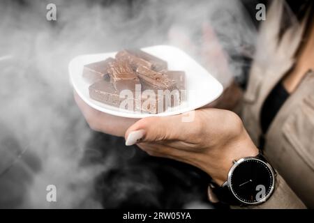 Das Mädchen zieht Aufmerksamkeit auf sich, indem es eine weiße Untertasse mit Schokoladenstücken im Rauch des Shisha-Nachtlebenskonzepts in der Hand hält. Stockfoto