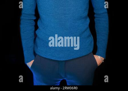 Schlanker Kerl in blauem Rollkragenkleid für Herren mit Händen in der Tasche auf schwarzem dunklem Hintergrund. Stockfoto