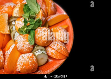 Obstteller mit Mandarine, Birne, Persimmon und mit Minzblättern garniert und auf dem Hintergrund schwarz mit Puderzucker bestreut. Stockfoto