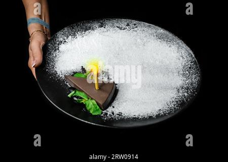 Die Hand einer Frau hält einen dunklen Teller mit einem süßen Schokoladendessert, Käsekuchen und einer brennenden Weihnachtskerze und weißem Puderzucker auf einem schwarzen Backgrou Stockfoto