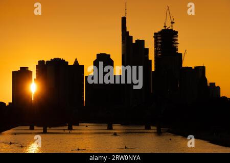 Die Sonne scheint zwischen den Wolkenkratzern der Frankfurter Bank-Skyline kurz vor dem Untergang, während einige Ruderer und Stand-up-Paddler Stockfoto