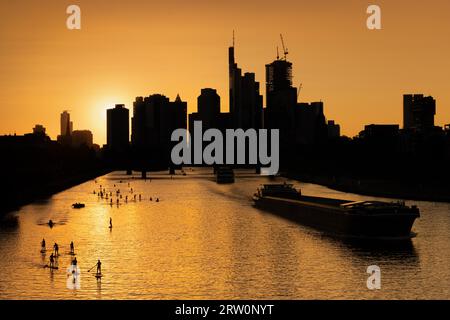 Die Sonne scheint zwischen den Wolkenkratzern der Frankfurter Bank-Skyline kurz vor dem Untergang, während einige Schiffe, Boote, Ruderer und Stockfoto