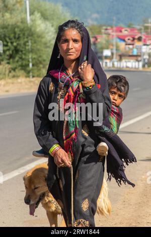 Anantnag, Indien. September 2023. Eine Kaschmir-Nomadenfrau, die vor dem Winter ein Kind auf dem Rücken in wärmere Ebenen trägt, in der Gegend von Kokernag, etwa 100 km von Srinagar entfernt. Jedes Jahr reisen Tausende nomadischer Bakerwal- und Gujjar-Familien in die Hochwiesen Kaschmirs und bleiben dort für die Sommermonate, um Vieh zu weiden. Sie fahren vor dem Winter im Oktober zurück in die wärmeren Ebenen. Quelle: SOPA Images Limited/Alamy Live News Stockfoto