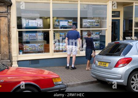 Leute, die Häuser zum Verkauf in einem Immobilienmakler-Fenster am 13. September 2023 in Cirencester, Großbritannien, suchen. Cirencester ist bekannt dafür, dass es in seinem kleinen Stadtzentrum eine große Anzahl von Immobilienmaklern gibt, da der ländliche Wohnungsmarkt weiterhin aktiv ist. Cirencester ist eine Marktgemeinde in Gloucestershire. Es ist die achtgrößte Siedlung in Gloucestershire und die größte Stadt innerhalb der Cotswolds. Die meisten Gebäude und Häuser in der Stadt sind aus dem charakteristischen honigfarbenen Cotswold Stone gefertigt. Stockfoto