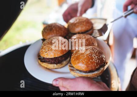 Prozess des Kochens und Grillens von Rindfleischburgern auf dem Open-Air-Straßenfest, Blick auf die Hände des Küchenchefs in Handschuhen mit verschiedenen Zutaten auf einem Grill Stockfoto