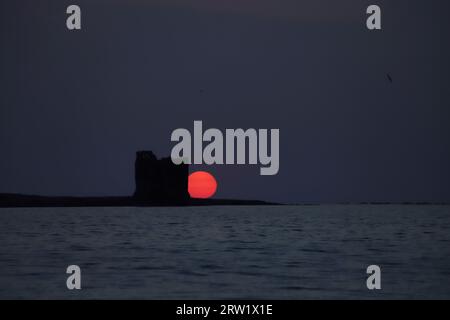 Eine lebhafte Orange Blood Sun, die hinter der Silhouette des Torre della Finanza auf Sardinien untergeht. Vom Fornelli-Kanal aus gesehen, mit Blick auf das Meer bei Sonnenuntergang. Stockfoto