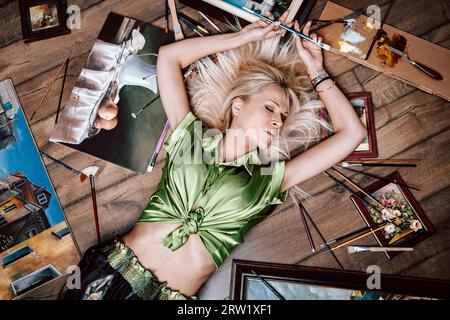 Blick von oben auf eine junge blonde Malerin, umgeben von ihren Kunstwerken Stockfoto
