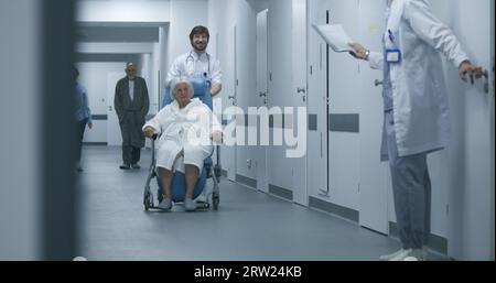 Der Arzt schiebt den Rollstuhl mit der Patientin, die durch den Klinikkorridor geht. Reife Ärztin öffnet Tür, eine andere bringt ältere Frau in den OP. Medizinisches Personal und Patienten im Spitalgang. Stockfoto