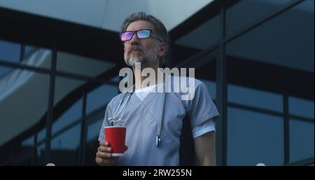Reifer Arzt in Brille und Uniform steht am Abend in der Nähe des Klinikeingangs. Professioneller Sanitäter trinkt Kaffee. Medizinisches Personal des Krankenhauses oder des modernen medizinischen Zentrums. Lifestyle- und Gesundheitskonzept. Stockfoto