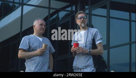 Zwei Ärzte in Uniform stehen am Abend in der Nähe des Eingangs zum medizinischen Zentrum. Professionelle Sanitäter trinken Kaffee, diskutieren Arbeit. Medizinisches Personal des Krankenhauses oder der modernen Klinik. Lifestyle- und Gesundheitskonzept. Stockfoto