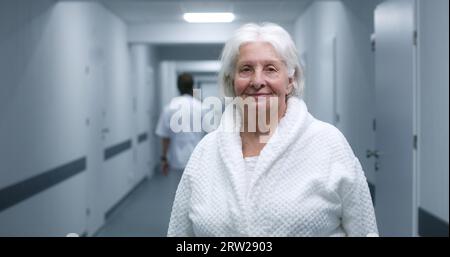 Die Seniorin steht in einem modernen Klinikkorridor. Ältere Patientin lächelt und schaut in die Kamera. Medizinisches Personal, Ärzte und Patienten im Hintergrund im Krankenhaus oder medizinischen Zentrum Flur. Hochformat. Stockfoto