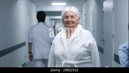 Die Seniorin steht in einem modernen Klinikkorridor. Ältere Patientin lächelt und schaut in die Kamera. Medizinisches Personal, Ärzte und Patienten im Hintergrund im Krankenhaus oder medizinischen Zentrum Flur. Hochformat. Stockfoto